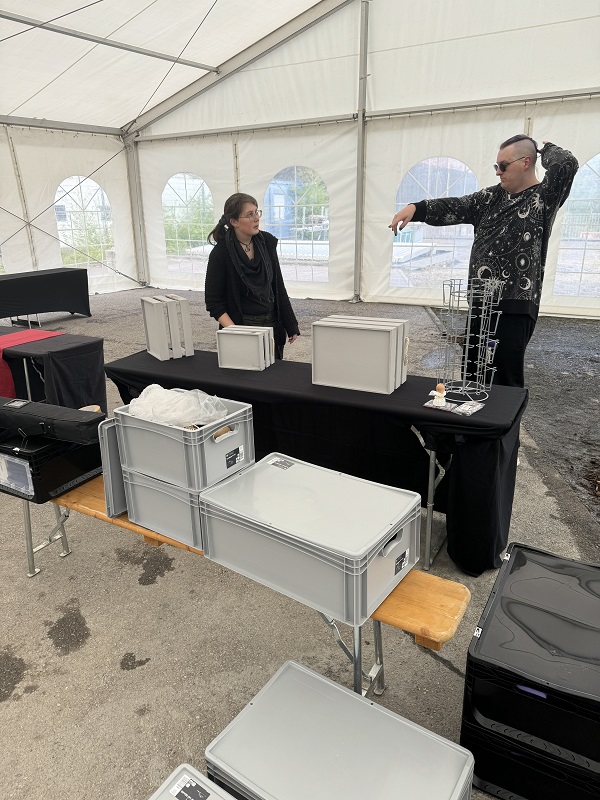 Zwei Personen stehen in einem großen Veranstaltungszelt und beraten darüber, wie ein Verkaufsstand aufgebaut werden soll, es sind verschiedene Tische und Boxen zu sehen